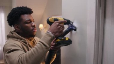 African American man in hoodie drills hole in concrete wall with hammer drill. Man does repairs in his apartment. Repair work, installation of the Internet or making hole for the picture. Dolly shot.