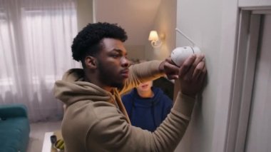 African American man connects security camera with wire and fastening on wall. Multi ethnic couple installs cameras in their apartment. Concept of CCTV cameras, monitoring system, safety and privacy.