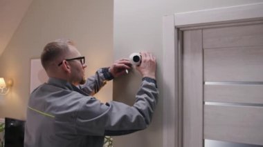 Installer in uniform puts security camera on wall fastening and connects it to system with cable. Man installs cameras in house. Concept of CCTV cameras, monitoring, safety and privacy. Zoom in shot.