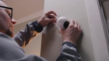 Installer in uniform puts security camera on wall mount and connects it to system with wire. Man installs cameras in apartment. Concept of CCTV cameras, tracking, safety and privacy. Zoom in shot.