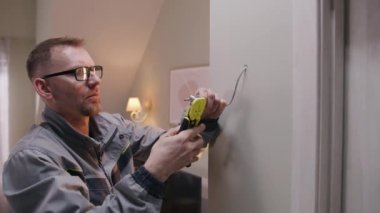 Man in uniform and glasses works with cable in drilled hole. Electrician removes insulation from wire with professional tool in the apartment. Installation of Internet or electricity work. Zoom in.