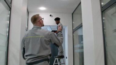 Two multi ethnic installers in uniform sets up security cameras in office corridor using laptop. Upset African American worker looks at camera. Concept of surveillance systems and privacy. Quick zoom.