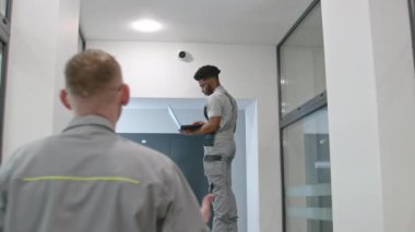 Two multi ethnic installers uniform sets up security cameras in office corridor using laptop. Upset African American worker looks at camera. Concept of surveillance systems and privacy. Quick zoom in