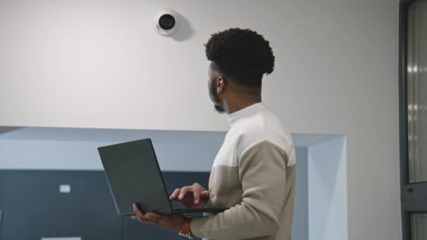 アフリカ系アメリカ人のインストーラは ラップトップを使用してオフィスルームにセキュリティカメラを設定します 男はコンピュータープログラムでCctvカメラをチェックしてる 監視および追跡システム 監視システムとプライバシーの概念 — ストック動画