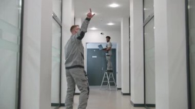 Two multi ethnic installers in uniform sets up security cameras in office corridor using laptop. They talk, check CCTV cameras in computer program. Concept of monitoring system and privacy. Dolly shot