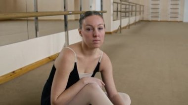 Female ballet dancer after performance choreography rehearsal. Exhausted ballerina in training bodysuit sits on floor after gymnastic workout in dance studio, looks at camera. School of ballet dance.