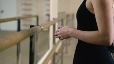 Ballerina in training bodysuit stands near ballet barre in dance studio and prepares for stretching before performance. Ballet dancer on choreography rehearsal in dance hall. Ballet dance school.