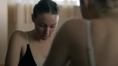 Upset ballerina in training bodysuit stands near ballet barre in dance studio and looks at herself in mirror. Female ballet dancer before stretching and gymnastic workout. Classic ballet dance school.