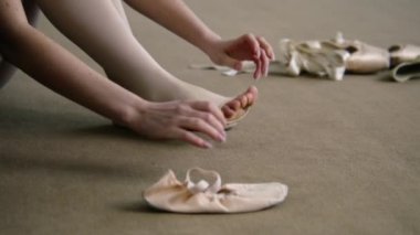 Beautiful ballet dancer sits on floor near pointe at dance studio and puts on ballet shoes before choreography lesson. Ballerina prepares for performance. Classical ballet dance school. Feet close up.