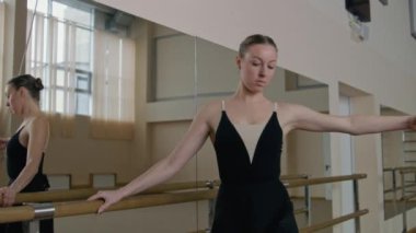 Female ballet dancer on choreography rehearsal in dance studio with mirrors. Adult ballerina in training bodysuit rehearses near ballet barre and practices classical ballet movements. Dance school.