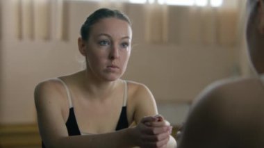 Tired ballerina in training bodysuit stands near ballet barre in dance studio and looks at herself in mirror. Female ballet dancer before stretching and gymnastic workout. Classic ballet dance school.