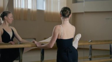 Female ballet dancer doing gymnastic exercises in front of the mirror. Ballerina in training bodysuit stands near ballet barre in dance studio, stretches her leg and warms up. School of ballet dance.
