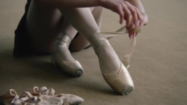 Adult ballerina sits on floor at dance studio and puts on pointe shoes before choreography lesson. Beautiful dancer prepares for performance or training. Classical ballet dance school. Feet close up.