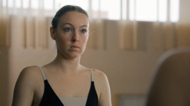 Tired ballerina in training bodysuit stands near ballet barre in dance studio and looks at herself in mirror. Female ballet dancer before stretching and gymnastic workout. Classic ballet dance school.