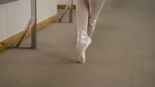 Close-up feet shooting of professional ballerina standing on tiptoe in pointe shoes at dance studio. Ballet dancer prepares for performance. Woman doing classic ballet exercises. Classic ballet school