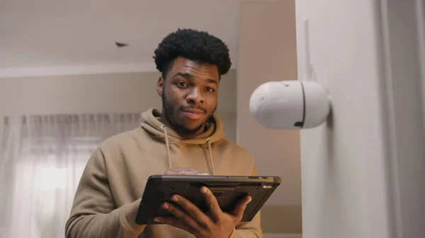 African American man installs security camera. Man sets up angle of CCTV camera at home and rotates it with digital tablet and application. Monitoring system, safety and privacy concept.