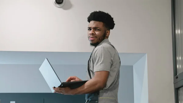 Two multi ethnic installers in uniform sets up security cameras in office corridor using laptop. Upset African American worker looks at camera. Concept of surveillance systems and privacy.