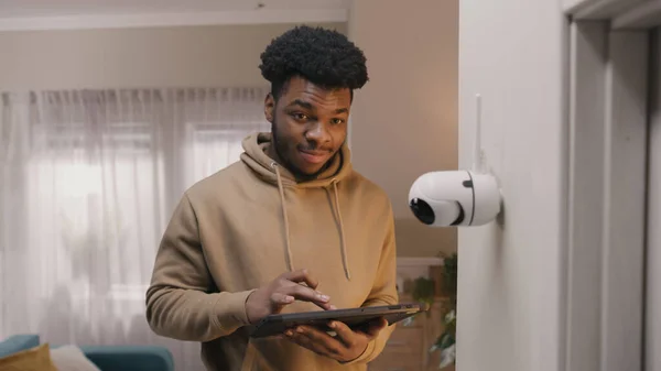 African American man installs security camera. Man sets up angle of CCTV camera at home and rotates it using digital tablet and application. Monitoring system, safety and privacy concept.