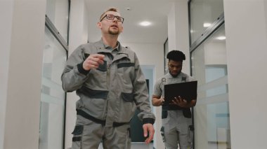 Two diverse installers in uniform walk hallway and discuss security cameras installation in modern college. Men set up CCTV cameras using computer software on laptop. Concept of surveillance system.