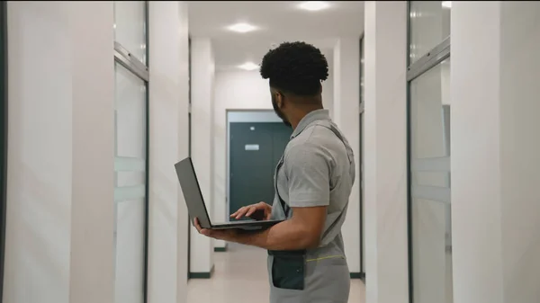African American installer in uniform stands in hallway and sets up security cameras using program on laptop. CCTV cameras installation in coworking office. Concept of surveillance system.
