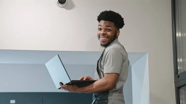 Male installer in uniform sets up security camera in office corridor using laptop. Funny African American worker looks at camera and smiles. Concept of surveillance and monitoring systems.