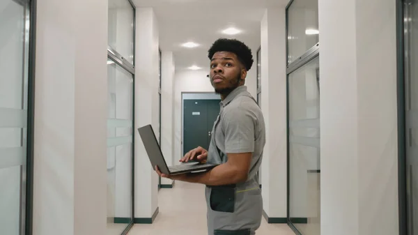 African American installer in uniform stands in hallway and sets up security cameras using program on laptop. CCTV cameras installation in coworking office. Concept of surveillance system.
