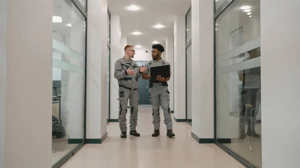 Two diverse men in uniform walk corridor and discuss CCTV cameras installation in coworking office. Installers set up security cameras using computer program on laptop. Concept of surveillance system.