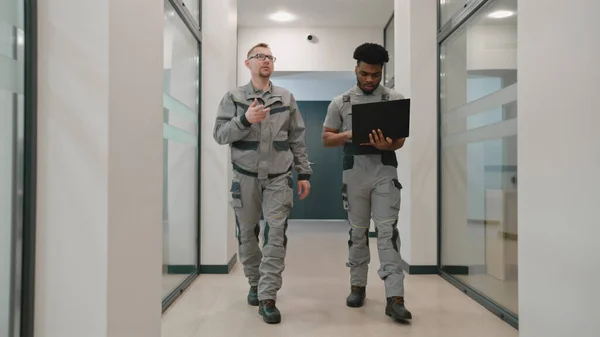 Two diverse men in uniform walk corridor and discuss CCTV cameras installation in coworking office. Installers set up security cameras using computer program on laptop. Concept of surveillance system.