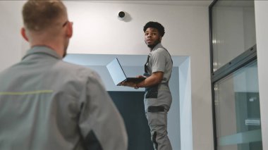 Two multi ethnic installers in uniform sets up security camera in office hallway using laptop. They talk, check CCTV cameras in computer program. Concept of surveillance system and privacy. Dolly shot