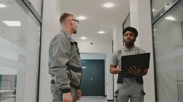 Two diverse installers in uniform walk hallway and discuss security cameras installation in modern college. Men set up CCTV cameras using computer software on laptop. Concept of surveillance system.