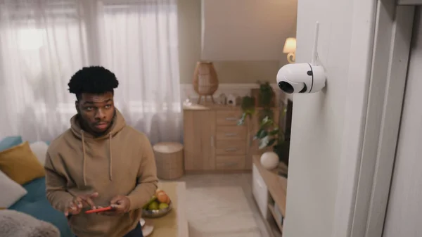 African American man completes installation of security camera. Man sets up angle of CCTV camera at home and rotates it using smartphone and app. Concept of monitoring system, safety and privacy.