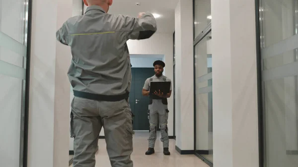 Two diverse men in uniform walk corridor and discuss CCTV cameras installation in business office. Installers set up security cameras using computer program on laptop. Concept of surveillance system.