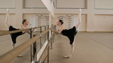 Graceful ballerina in training bodysuit stands near ballet barre, stretches leg and body, prepares for performance. Adult woman doing gymnastic exercises in dance studio. Classic ballet dance school.