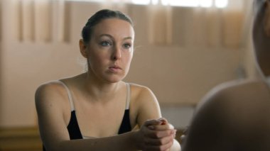 Tired ballerina in training bodysuit stands near ballet barre in dance studio and looks at herself in mirror. Female ballet dancer before stretching and gymnastic workout. Classic ballet dance school.