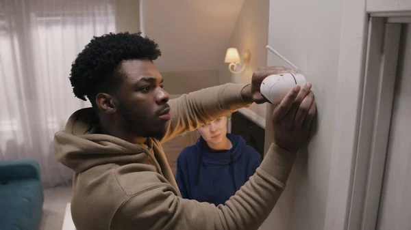 African American man connects security camera with wire and fastening on wall. Multi ethnic couple installs cameras in their apartment. Concept of CCTV cameras, monitoring system, safety and privacy.