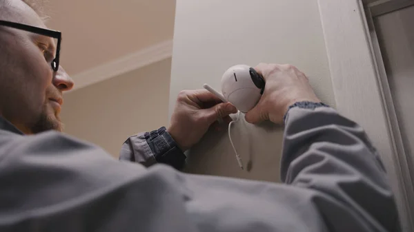 stock image Installer in uniform puts security camera on wall fastening and connects it to system with cable. Man installs cameras in house. Concept of CCTV cameras, monitoring, safety and privacy.