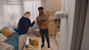 Multi ethnic couple install and set up security camera. Woman moves to check motion sensor on CCTV camera, man uses digital tablet and application. Concept of tracking system, safety and privacy.