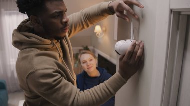 African American man connects security camera with wire and fastening on wall. Multi ethnic couple installs cameras in their apartment. Concept of CCTV cameras, monitoring system, safety and privacy. clipart