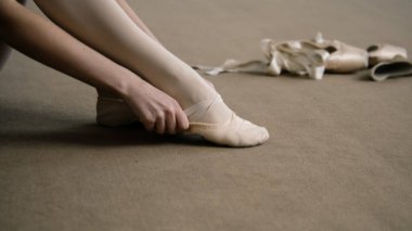 Ballerina sits on floor at dance studio and puts on pointe shoes before choreography lesson. Graceful ballet dancer prepares for performance or training. Classical ballet dance school. Feet close up.