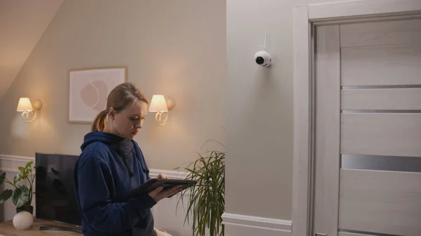Caucasian woman completes installation of security camera. Woman sets up CCTV camera at home using digital tablet computer and application. Monitoring and tracking system, safety and privacy concept.