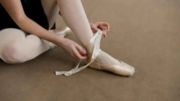 Ballerina in training bodysuit takes off her pointe shoes after stretching and gymnastic workout in dance studio. Female ballet dancer finished preparing for performance. Classic ballet dance school.