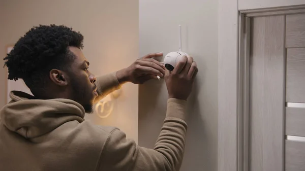 African American man puts security camera on wall fastening and connects it to system with cable. Man installs cameras in his apartment. Concept of CCTV cameras, tracking system, safety and privacy.