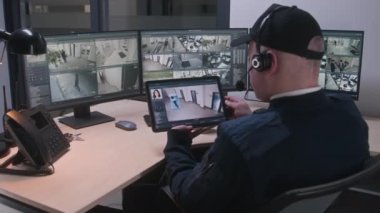 Kulaklıktaki güvenlik görevlisi, çalışma ofisindeki kapalı devre kameraları kontrol ediyor ve izliyor. Dijital tablet ve ekranda oynatıcısı olan bilgisayarlar kullanıyor. Güvenlik kamerası görüntülerini gösteren yüksek teknoloji yazılımı.