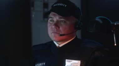 Güvenlik görevlisi işyerinde oturuyor ve monitörde gösterilen güvenlik kameralarına bakıyor. Adam iş arkadaşıyla telsizle konuşuyor. İzleme sistemi ve CCTV. Geç saatlere kadar çalışıyorum. Dikey görünüm.