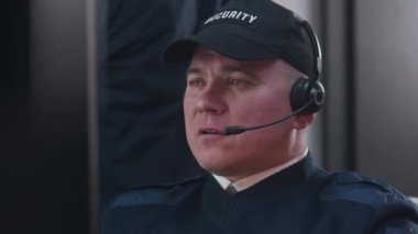 Kulaklıklı güvenlik görevlisi iş yerinde oturur ve monitörde gösterilen güvenlik kamerasından görüntüye bakar. Adam iş arkadaşıyla telsizle konuşuyor. İzleme sistemi ve CCTV. Dikey görünüm.