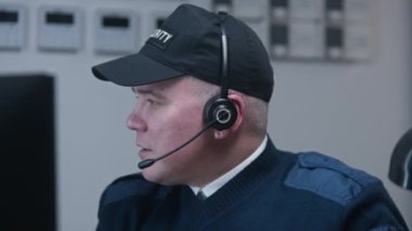 Üniformalı bir güvenlik görevlisi masanın yanında oturur ve monitörde gösterilen güvenlik kamerasından görüntüye bakar. Kulaklıklı adam iş arkadaşıyla konuşuyor. Gözlem ve CCTV teknolojisi. Dikey görünüm.