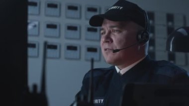 Güvenlik görevlisi masanın başında oturur, güvenlik kameralarından bilgisayar monitörüne bakar ve iş arkadaşıyla telsizle konuşur. İzleme ve izleme sistemi. CCTV teknolojisi. Dikey görünüm