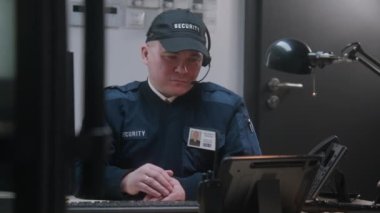 Kulaklıklı bir güvenlik çalışanı iş yerinde oturuyor, güvenlik kamerasından monitöre ve klavyeye bakıyor. Adam operatörü tabletle arıyor. İzleme ve izleme sistemleri. CCTV teknolojisi.