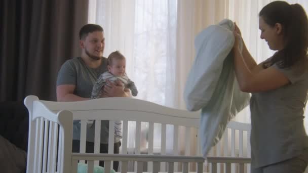 母親はベビーベッドに毛布を置き 赤ちゃんを見て話します 子育て中の父親は 赤ん坊の娘や息子を腕の中に抱えており ケーブルの準備を見ています 幼少期 親子の概念 — ストック動画