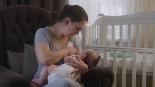 母親の母乳育児小さな新生児 ベビーベッドの近くのアームチェアに座っています 母乳を供給しながら 女性は彼女の腕の中で赤ちゃんの娘や息子を保持しています 幼少期 母子家庭の概念 — ストック動画